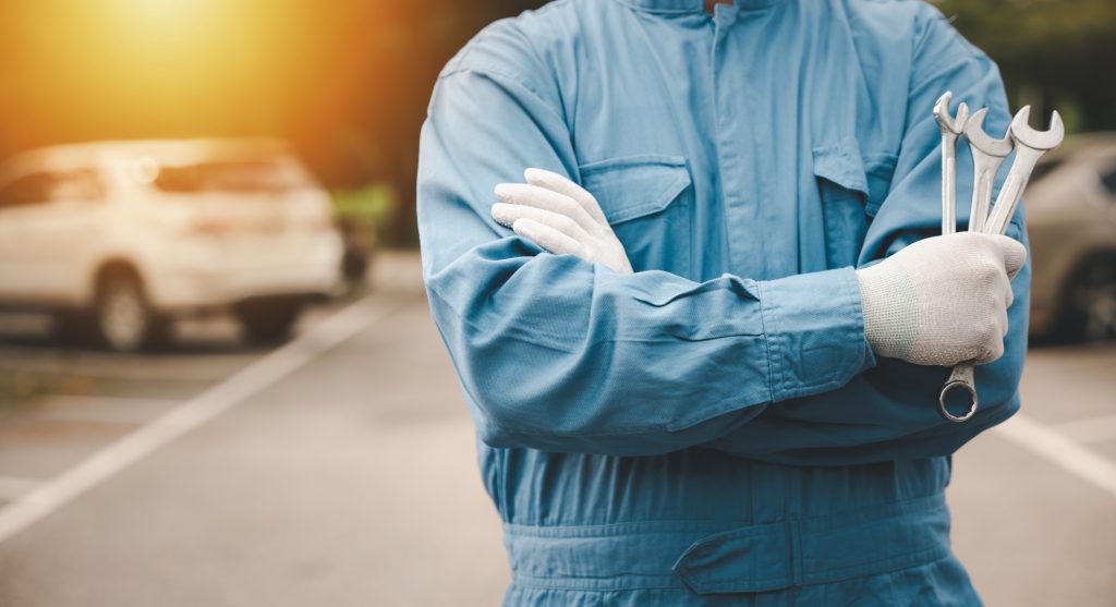 A mechanic wearing a blue jumpsuit and gloves, holding a set of wrenches with arms crossed, standing in a parking lot with a car in the background.